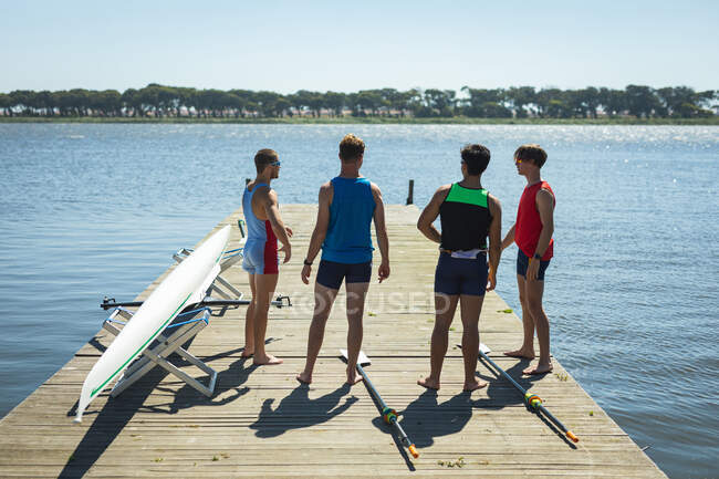 Vista posteriore di quattro rematori maschi caucasici in piedi scalzi su un molo, con remi sdraiati e una barca a remi su un molo — Foto stock