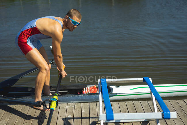 Side view of a Caucasian male rower preparing a rowing boat before rowing, standing on a jetty on the river holding an oar — Stock Photo