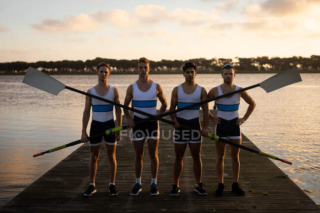 Portrait of four fit confident Caucasian male rowers, standing on a jetty on the river holding oars and looking to camera at sunset — Stock Photo