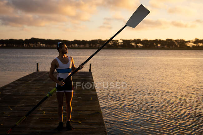 Front view of a confident fit Caucasian male rower standing on a jetty on the river, holding an oar on a sunny day at sunset — Stock Photo