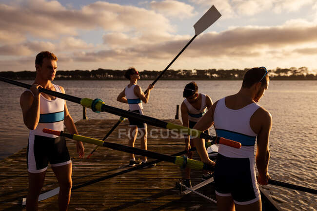 Front view of a rowing team of four Caucasian men standing on a jetty on the river, holding oars, one of them on his shoulders — Stock Photo