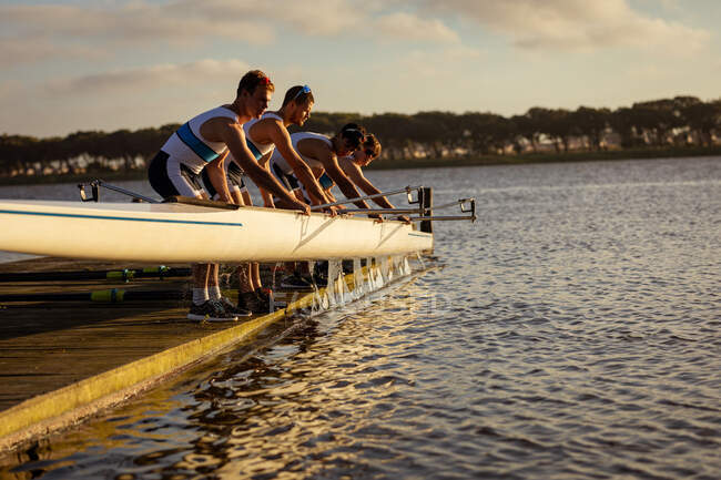 Side view of a rowing team of four Caucasian men taking a boat out of water standing on a jetty on the river at sunset — Stock Photo
