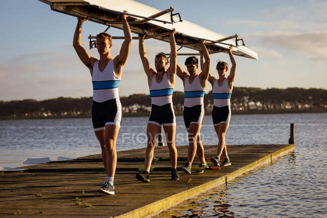 Vista frontal de un equipo de remo de cuatro hombres caucásicos llevando un bote sobre sus cabezas con los brazos levantados, caminando a lo largo de un embarcadero en el río al atardecer - foto de stock