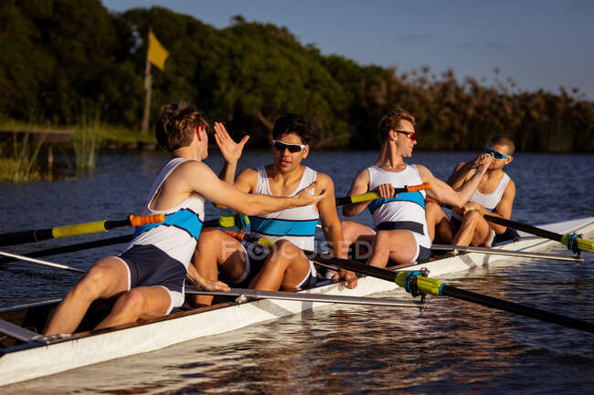 Vista frontal de uma equipe de remo de quatro homens caucasianos treinando e remando no rio, sentados em um barco a remo, de mãos dadas, curtindo o treinamento — Fotografia de Stock