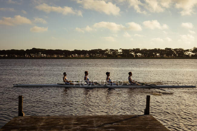 Біля входу в веслувальну команду з чотирьох кавказьких чоловіків, які тренуються і веслують на річці, сидячи в веслувальному човні на заході сонця. — стокове фото