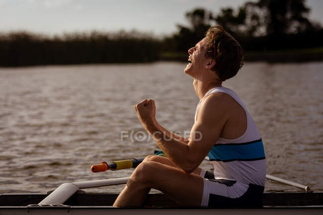 Vue latérale d'un rameur caucasien s'entraînant et ramant sur la rivière, assis dans un bateau à rames et souriant et gesticulant de bonheur, profitant de l'entraînement — Photo de stock
