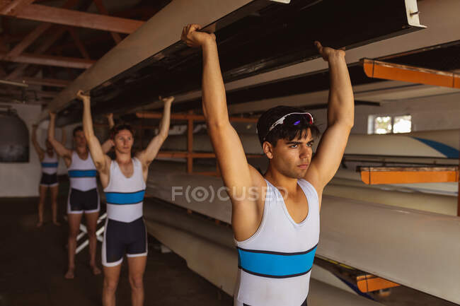 Front view of a rowing team of four Caucasian men carrying a boat above their heads with arms raised, walking from a boathouse to rowing practice — Stock Photo