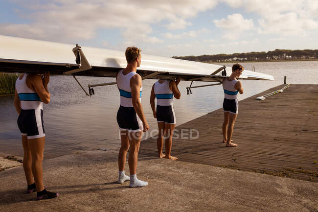 Rear view of a rowing team of four Caucasian men carrying a boat on their shoulders, walking along a jetty on the river in a sunny day — Stock Photo