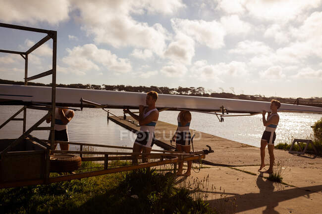 Side view of a rowing team of four Caucasian men carrying a boat on their shoulders, walking to a boathouse from a rowing practice — Stock Photo