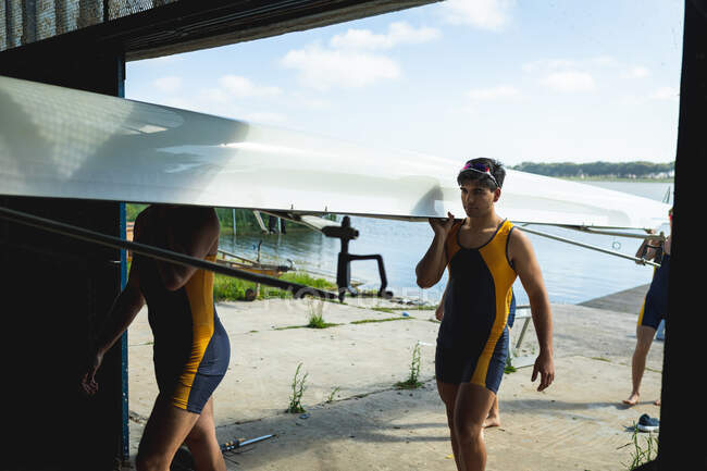 Vue de face d'une équipe d'aviron de quatre hommes caucasiens portant un bateau sur leurs épaules, marchant le long d'une jetée sur la rivière dans un hangar à bateaux — Photo de stock