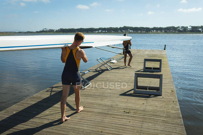Rear view of a rowing team of two Caucasian men carrying a boat on their shoulders, walking along a jetty on the river — Stock Photo