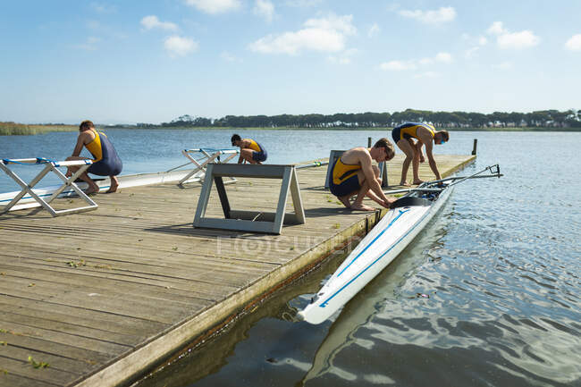 Vista lateral de un equipo de remo de cuatro hombres caucásicos poniendo botes de remo en el agua antes de remar, de pie en un embarcadero en el río en un día soleado - foto de stock
