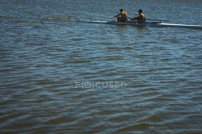 Вид сбоку гребной команды из двух кавказских мужчин, тренирующихся и гребящих на реке, держащих весла, сидящих в гребной лодке — стоковое фото