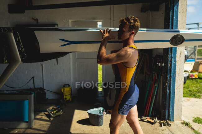 Seitenansicht eines kaukasischen Ruderers, der ein Boot auf seinen Schultern trägt und nach dem Rudern zu einem Bootshaus geht, an einem sonnigen Tag — Stockfoto