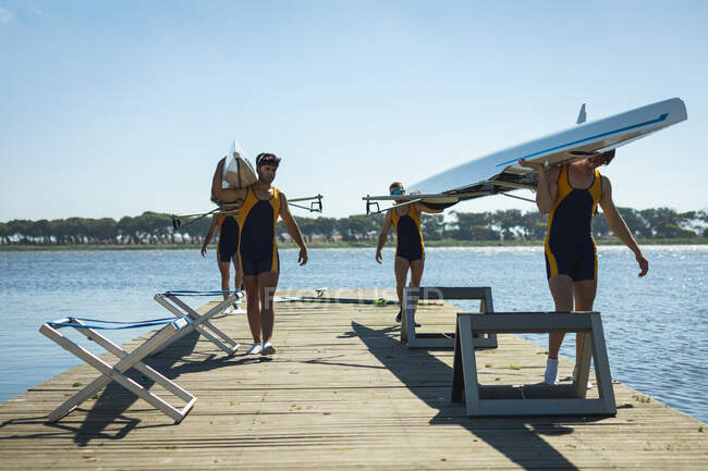 Перед ними вимальовується веслувальна команда з чотирьох кавказьких чоловіків, які носять човен на плечах, йдучи вздовж пристані на річці в сонячний день. — стокове фото