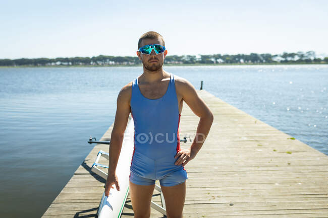 Передній вид кавказького веслувальника стоїть на пристані на річці в сонячний день, в сонцезахисних окулярах, дивлячись на камеру, з човном поруч. — стокове фото