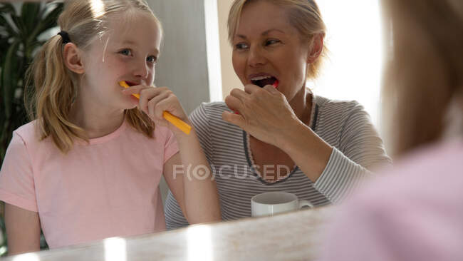 Por encima de la vista del hombro de una mujer caucásica disfrutando de tiempo en familia con su hija en casa juntos, cepillándose los dientes y sonriendo en un baño reflejado en el espejo - foto de stock