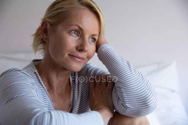 Vista frontal de una mujer caucásica disfrutando del tiempo libre en casa, sentada y pensando en su dormitorio, apoyada sobre su cabeza - foto de stock