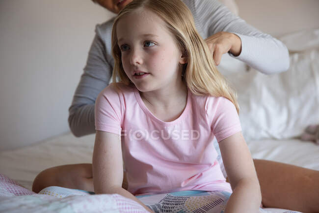 Передній вигляд поруч з кавказькою жінкою, яка проводить сімейний час зі своєю донькою вдома, мати чистить волосся своєї дочки сидячи на ліжку у своїй спальні. — стокове фото