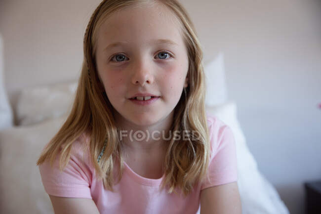 Portrait close up of a happy Caucasian girl enjoying free time at home, sitting on a bed in a bedroom, smiling and looking at camera, wearing pink t-shirt — Stock Photo