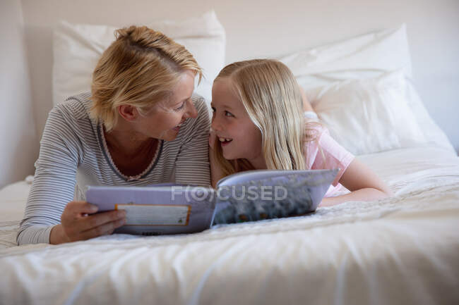 Вид спереди на кавказку, наслаждающуюся семейным отдыхом с дочерью дома вместе, читающую книгу и лежащую на кровати в своей спальне, улыбающуюся и смотрящую друг на друга — стоковое фото