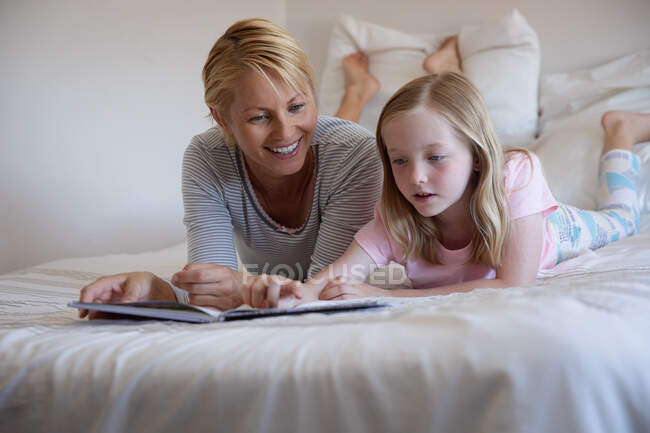 Vue de face d'une femme caucasienne profitant du temps en famille avec sa fille à la maison ensemble, lisant un livre et couché sur le lit dans leur chambre, souriant — Photo de stock