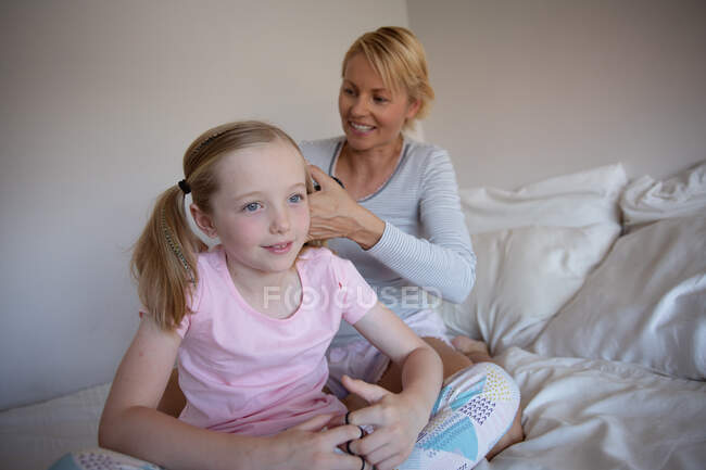 Vue de face d'une femme caucasienne profitant du temps en famille avec sa fille à la maison ensemble, la mère brossant les cheveux de sa fille, faisant queues de poney assis sur le lit dans leur chambre — Photo de stock