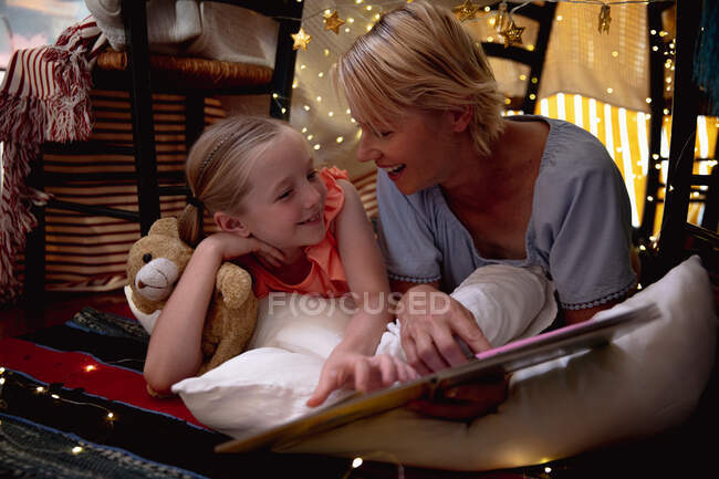 Front view of a Caucasian woman enjoying family time with her daughter at home together, smiling and talking in a tent in a sitting room, reading a book, with her daughter embracing her teddy bear — Stock Photo