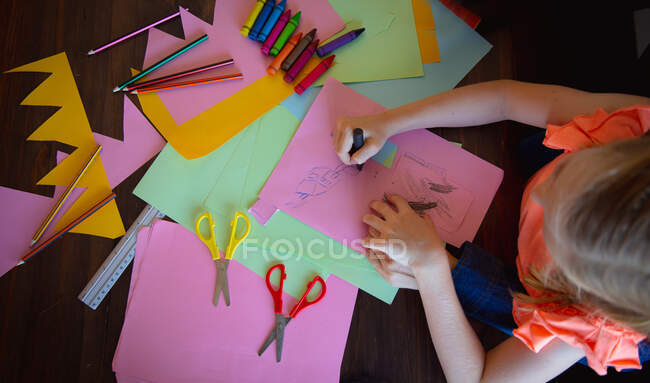 Visão aérea de uma menina caucasiana desfrutando de tempo livre em casa, sentada em uma mesa com tesoura, lápis na sala de estar e desenho em papéis coloridos — Fotografia de Stock