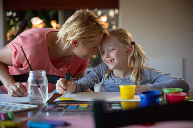 Vista laterale di una donna caucasica che si gode il tempo in famiglia con sua figlia a casa insieme, si siede a un tavolo in salotto, dipinge, sorride e si guarda — Foto stock