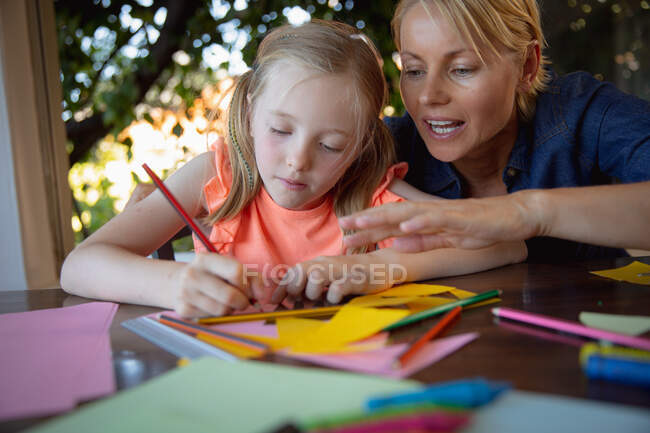 Vista frontal de uma mulher caucasiana desfrutando de tempo em família com sua filha em casa, sentada em uma mesa na sala de estar e desenhando papéis coloridos ajudando sua filha — Fotografia de Stock
