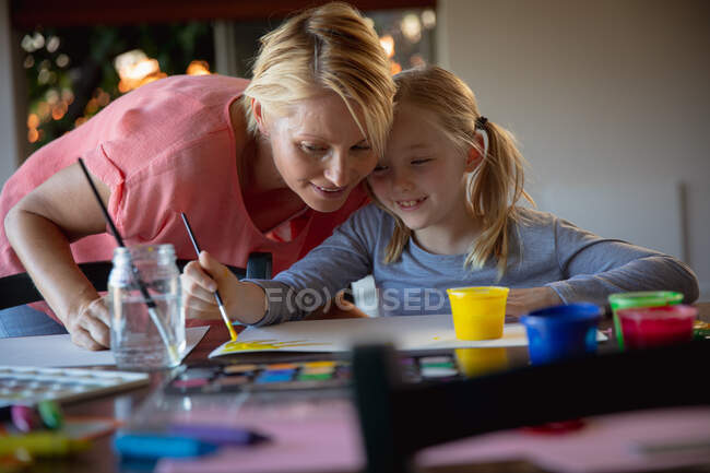 Вид спереди на кавказку, наслаждающуюся семейным отдыхом с дочерью дома, сидящую за столом в гостиной, рисующую и улыбающуюся — стоковое фото