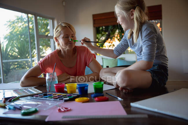 Vorderansicht einer kaukasischen Frau, die die Familienzeit mit ihrer Tochter zu Hause genießt, an einem Tisch im Wohnzimmer sitzt, malt und lächelt, die Tochter, die auf dem Tisch sitzt und die Nase ihrer Mutter bemalt — Stockfoto