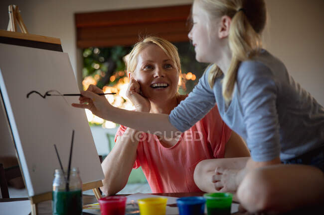 Front view of a Caucasian woman enjoying family time with her daughter at home together, sitting at a table in a sitting room, painting and smiling, the daughter sitting on a table, painting on canvas — Stock Photo