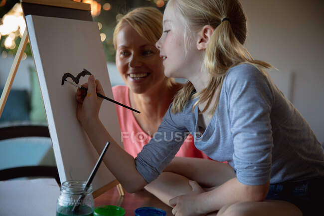 Vue de face d'une femme caucasienne profitant du temps en famille avec sa fille à la maison ensemble, assise à une table dans un salon, peignant et souriant, la fille assise sur une table, peinture sur toile — Photo de stock