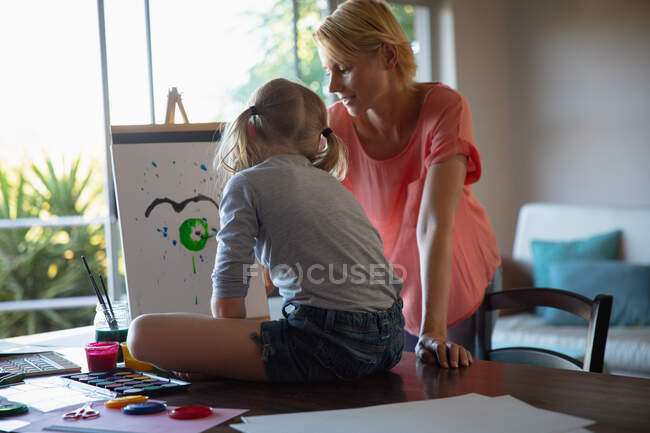 Vista laterale di una donna caucasica godendo di tempo in famiglia con la figlia a casa insieme, seduto a un tavolo in un salotto, pittura e sorridente, la figlia seduta su un tavolo, pittura su tela — Foto stock