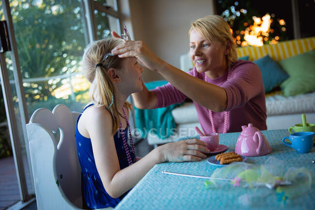 Vista lateral de uma mulher caucasiana desfrutando de tempo em família com sua filha em casa, colocando sobre ela a coroa, sorrindo e sentando-se a uma mesa na sala de estar — Fotografia de Stock