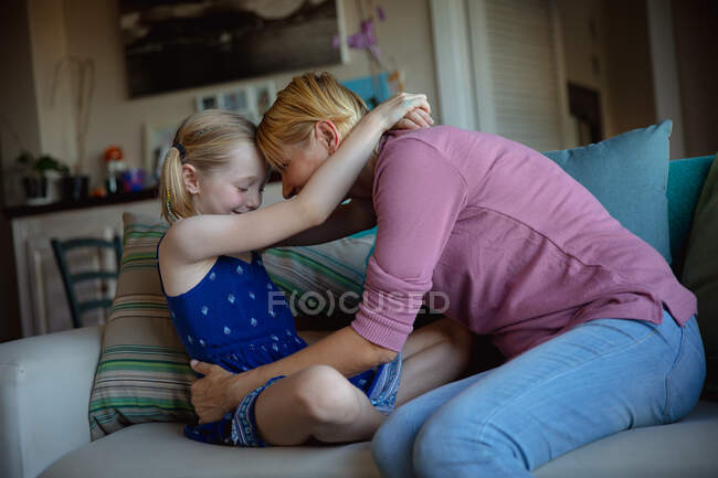 Side view of a Caucasian woman enjoying family time with her daughter at home together, sitting on a sofa in sitting room and embracing each other — Stock Photo