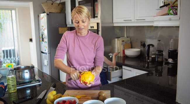 Vista frontal de una mujer caucásica disfrutando de tiempo libre en casa, pelando mango, preparando ensalada de frutas - foto de stock