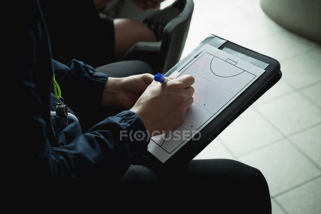 Side view mid section of male field hockey coach, sitting in the changing room, using a clipboard and drawing a game plan for the team — Stock Photo