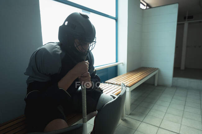 Seitenansicht eines kaukasischen männlichen Feldhockeyspielers, der in der Umkleidekabine sitzt, eine Gesichtsmaske trägt und einen Hockeyschläger hält, sich vor einem Spiel konzentriert und sein Kinn auf den Hokeystick lehnt — Stockfoto