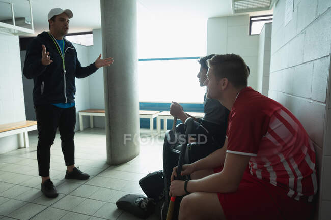 Сторона зору Кавказького тренера з хокею стоїть і взаємодіє з багатонаціональною групою хокеїстів-підлітків, сидячи в роздягальні, слухаючи його інструкції. — стокове фото