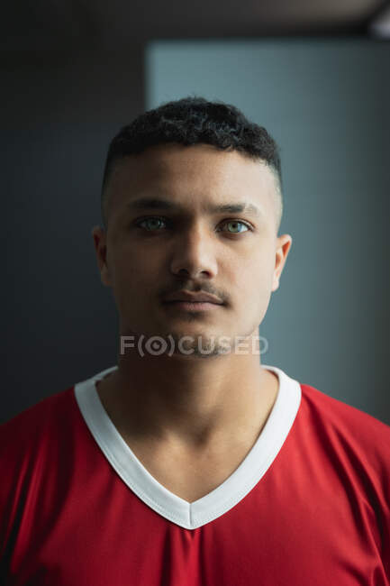 Portrait of a teenage mixed race male field hockey player, wearing a red  team strip, standing in a changing room looking straight to camera,  focusing before a game — focused, sportswear -