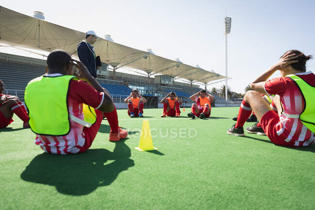 Low angle side view of a multi-ethnic group of teenage male field hockey players training before a game with their Caucasian male field hockey coach, working out on a field hockey pitch, doing sit ups in a circle on a sunny day — Stock Photo
