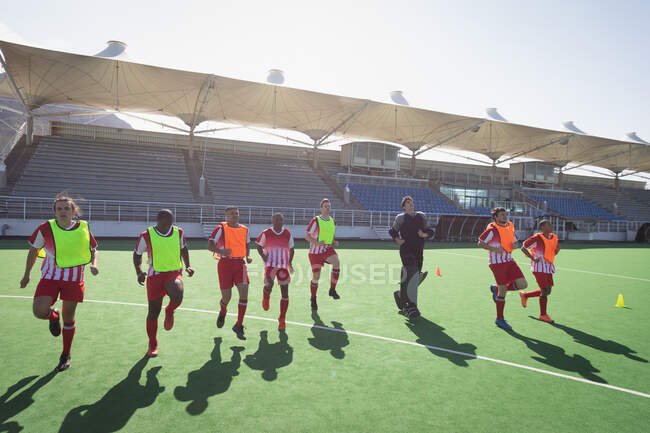 Side view of a multi-ethnic group of teenage male field hockey players, training before a game, working out with their coach on a field hockey pitch, running on a sunny day — Stock Photo