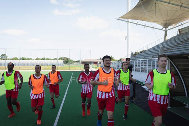 Front view of a multi-ethnic group of teenage male field hockey players, training before a game, working out on a field hockey pitch, running on a sunny day — Stock Photo