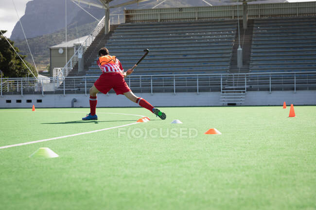 Rear view of a Caucasian male field hockey player, training before a game, hitting a ball with a hockey stick, with the stadium in the background on a sunny day — Stock Photo