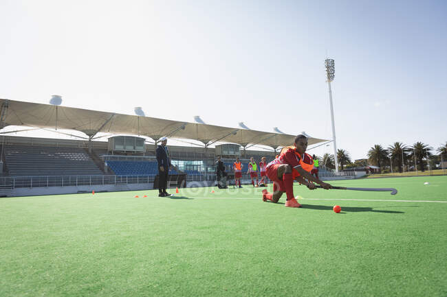 Side view of a mixed race male field hockey player, training on a field hockey pitch before a game, kneeling and trying to hit a ball with a hockey stick, with his teammates in the background on a sunny day — Stock Photo