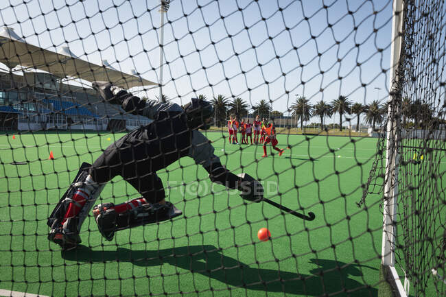 Rear view of a Caucasian male field hockey goalkeeper, diving to stop the ball with a hockey stick while training on the hockey pitch, with his teammates in the background on a sunny day — Stock Photo