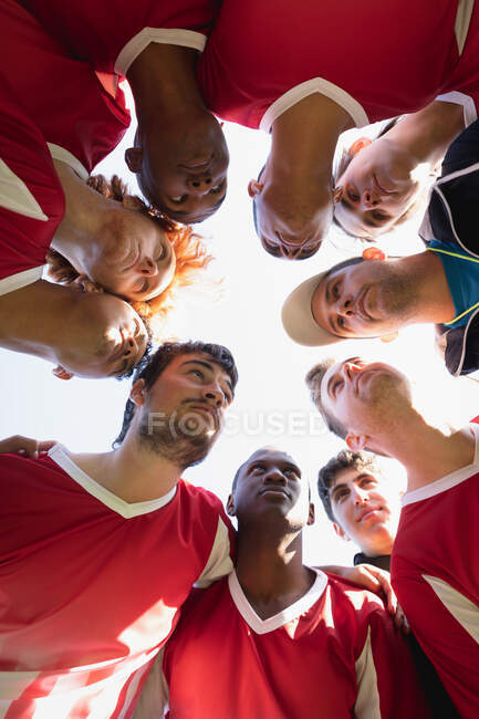 Visão de baixo ângulo, olhando para uma equipe multi-étnica de jogadores adolescentes de hóquei em campo e seu treinador de hóquei em campo masculino caucasiano se preparando antes de um jogo, em um huddle no campo, interagindo em um dia ensolarado — Fotografia de Stock